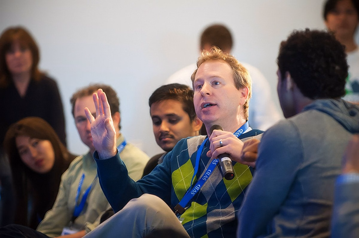 Man speaking into a microphone at a corporate event in Boston, with audience members visible in the background.