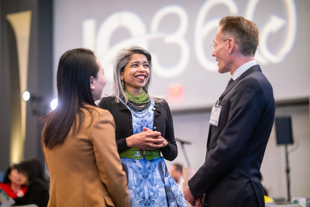 Three business professionals, two women and one man, engaging in a conversation at a conference or seminar, with a large digital clock in the background.
