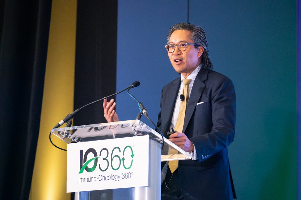A man in a suit and glasses speaking at a podium during a conference, with a sign that reads 'IP 360 Immuno-Oncology 360°'.
