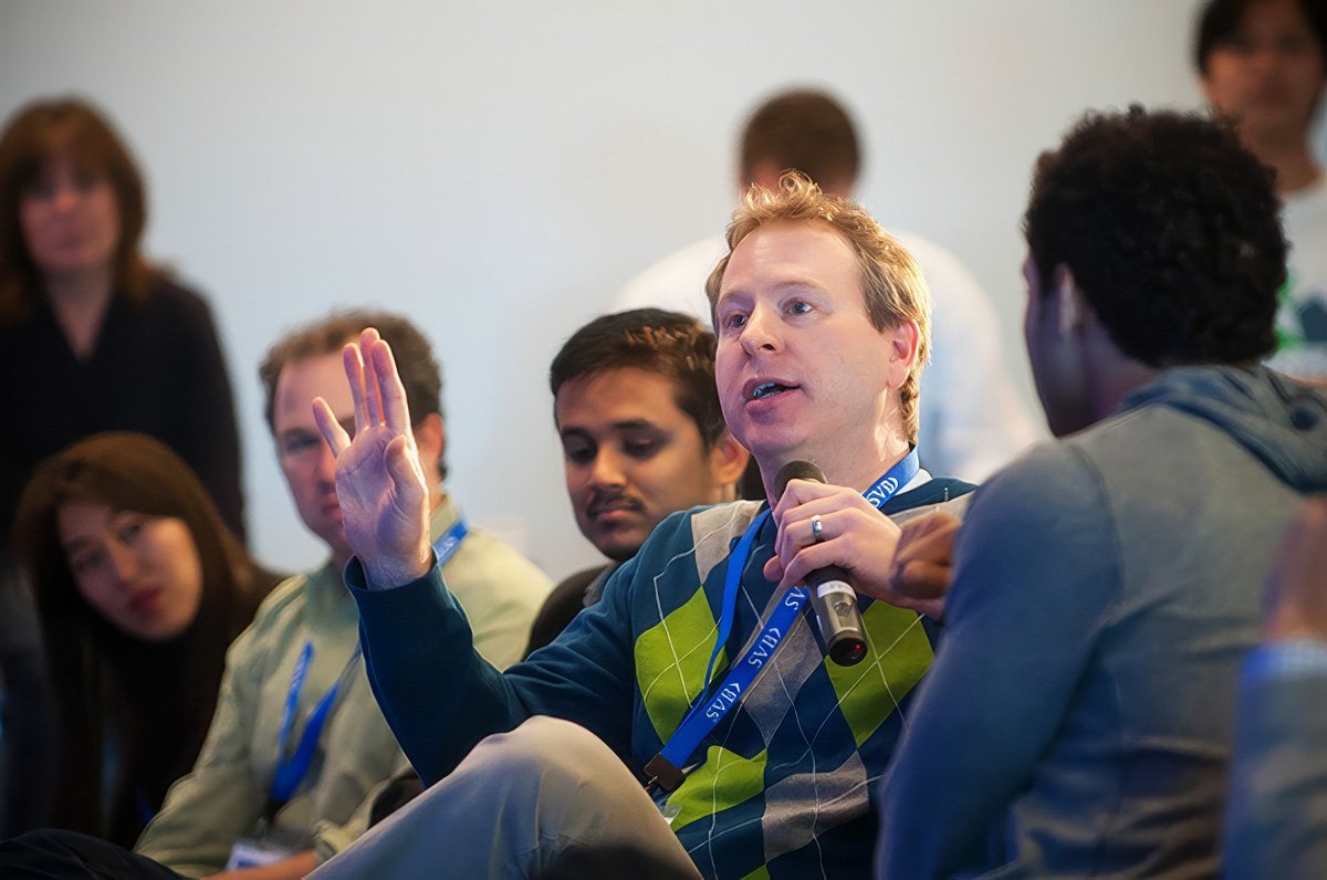 Man speaking into a microphone at a corporate event in Boston, with audience members visible in the background.