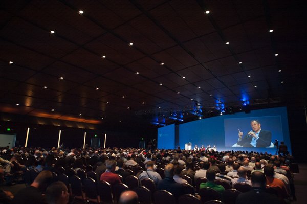 Large conference or seminar with many attendees, a big blue screen with a speaker on stage giving a presentation.