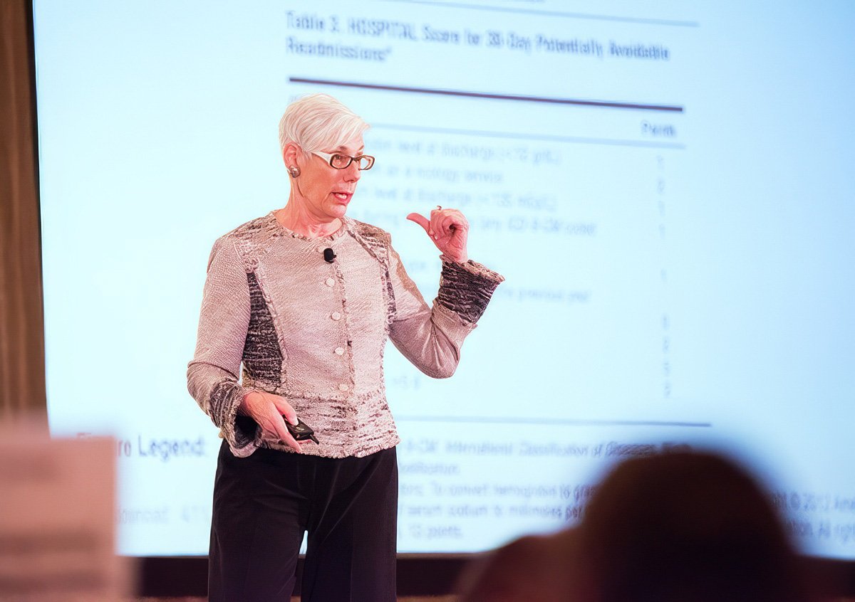 A woman in a stylish outfit presenting at a conference with a slide in the background.