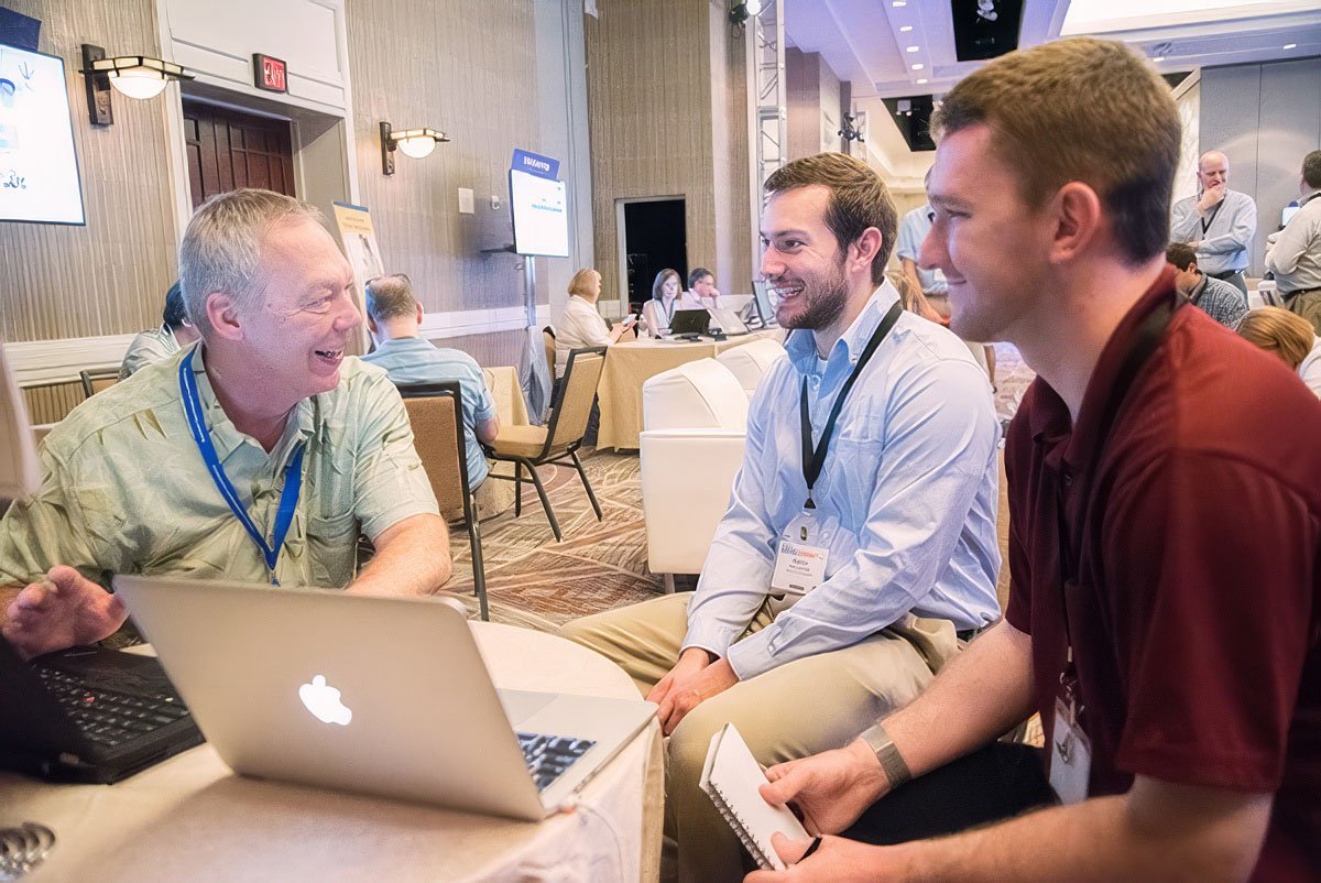 Three men engaged in a conversation at a corporate event, with laptops and notepads on the table.