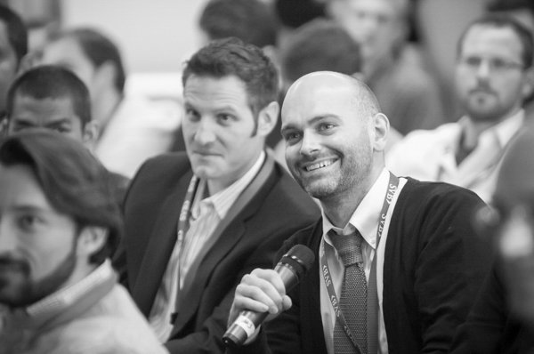 A man smiling and holding a microphone in a conference setting, with blurred attendees in the background.